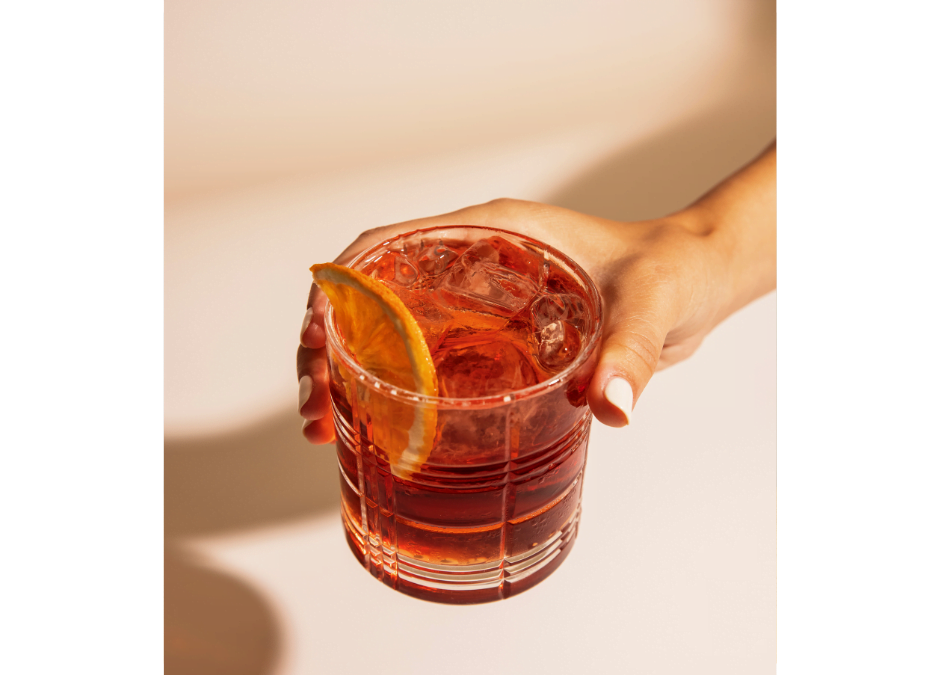 Sunshine Reggae alcoholvrije cocktail. Een hand heeft een tumblerglas vast, gevuld met de rood gekleurde sunshine reggae cocktail, ijsklontjes en afgewerkt met een schijfje sinaasappel